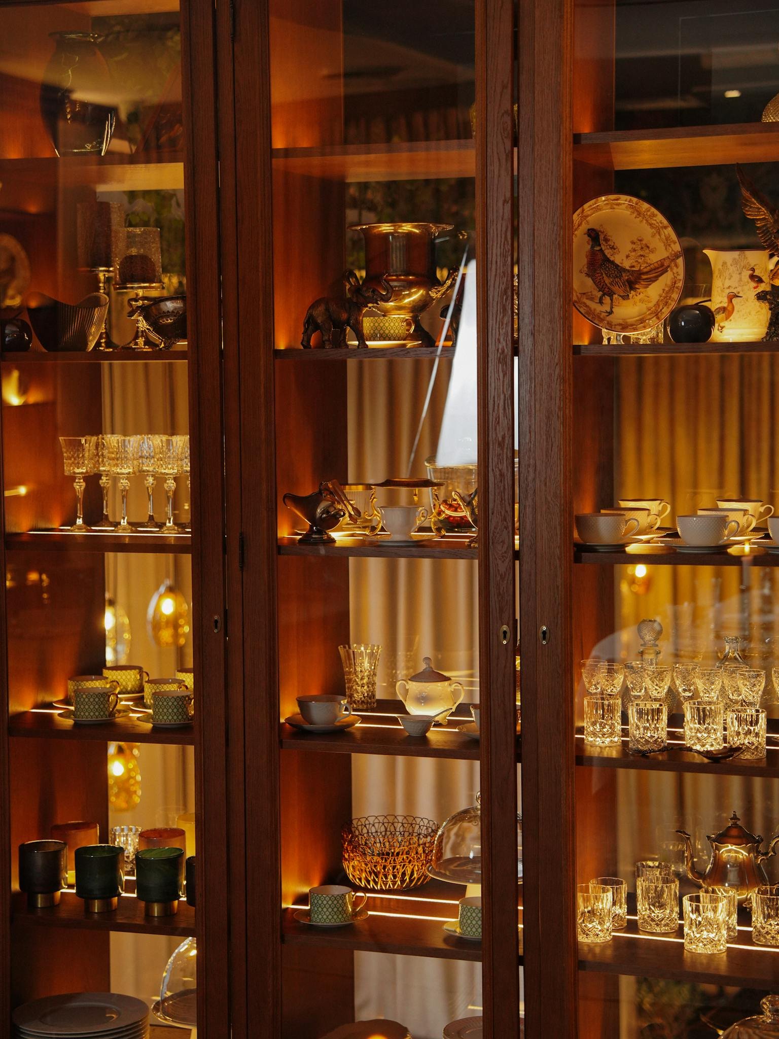 A sophisticated showcase of fine china, glassware, and home decor in a beautifully lit wooden cabinet.